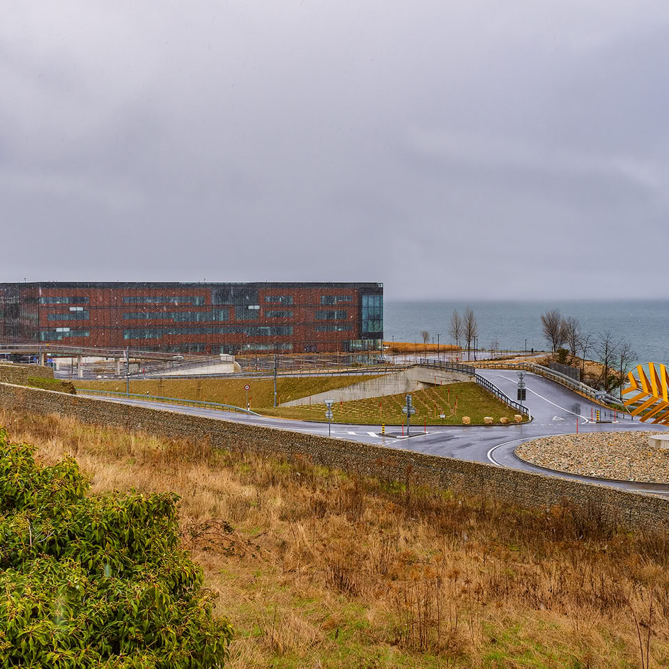 Paysage côtier avec un bâtiment moderne en arrière-plan, des arbres dénudés, un chemin sinueux, et la mer sous un ciel nuageux.