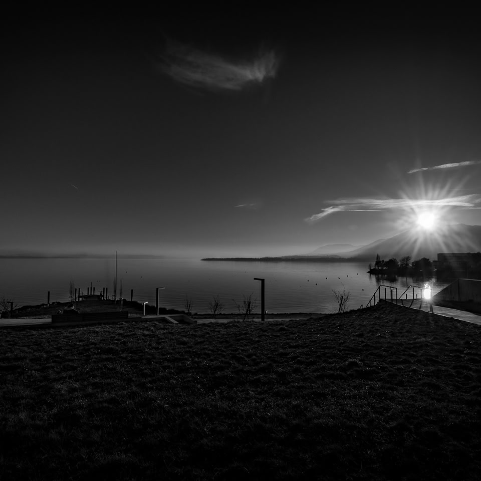 Paysage en noir et blanc avec un lac, un quai, des montagnes à l'horizon et le soleil se levant ou se couchant.