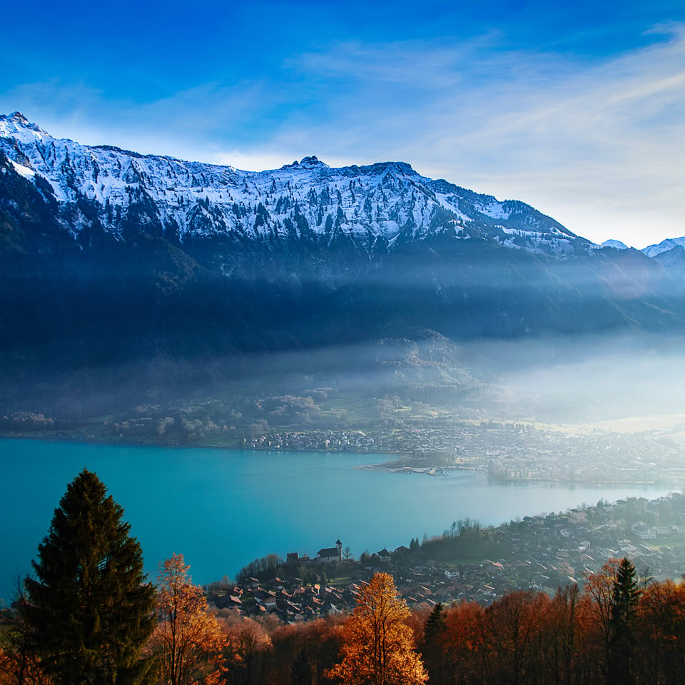 Paysage de montagne avec un lac turquoise au premier plan, des arbres aux couleurs automnales, et des montagnes enneigées à l'arrière-plan sous un ciel bleu.