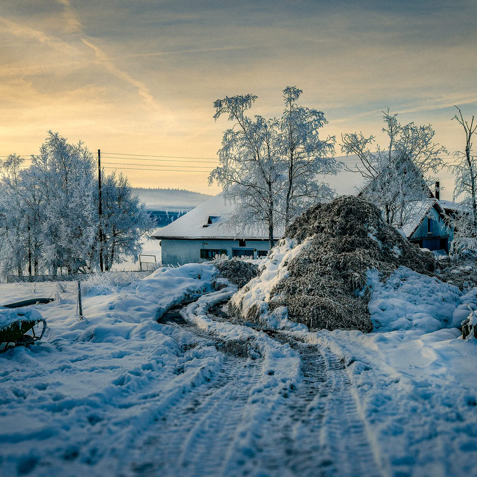 Paysage hivernal avec neige couvrant le sol, des arbres givrés, une maison et un tas de foin. Ciel partiellement nuageux au coucher du soleil.