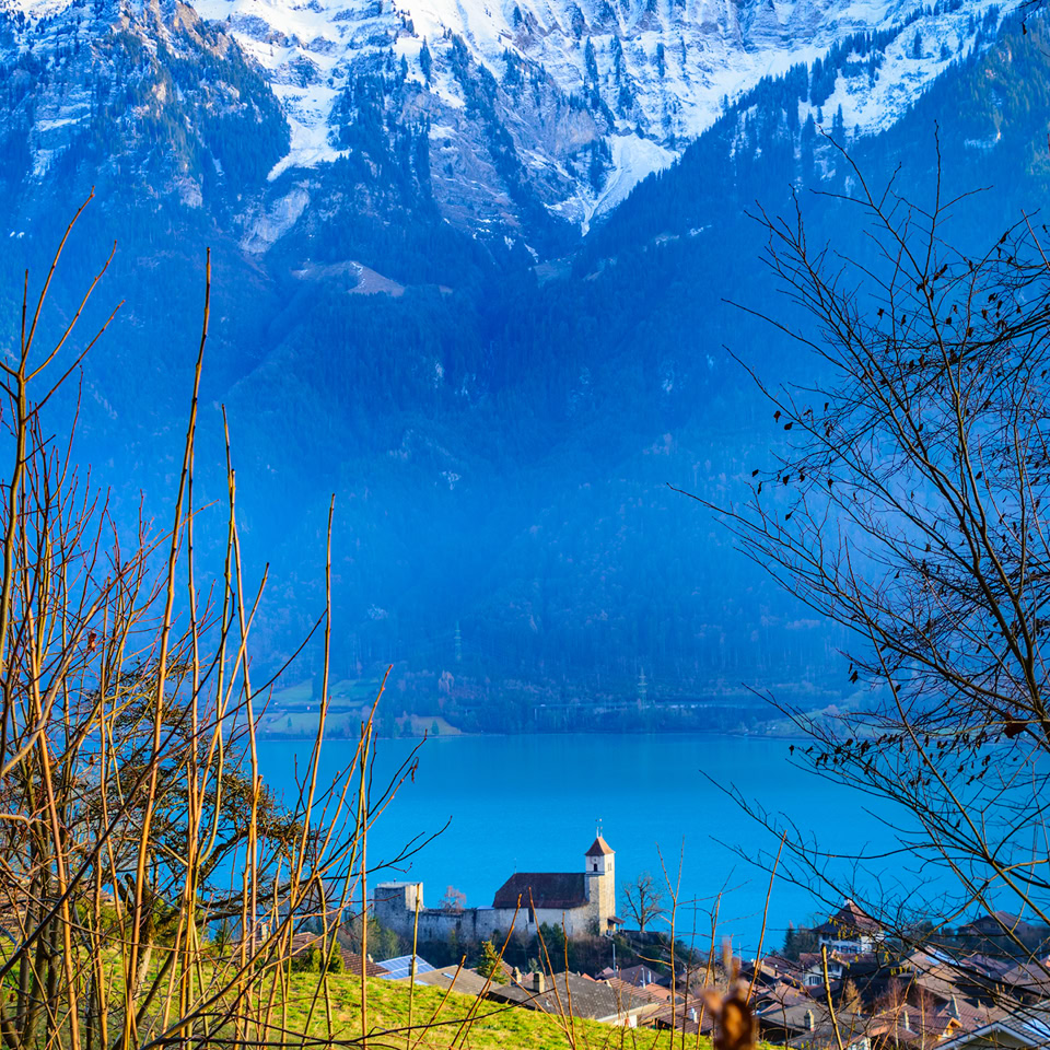 Paysage avec un lac, une église au bord de l'eau, des montagnes enneigées en arrière-plan et des arbres au premier plan.