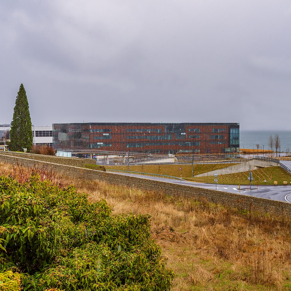 A modern building near a body of water, surrounded by grassy and landscaped areas, with a cloudy sky overhead.