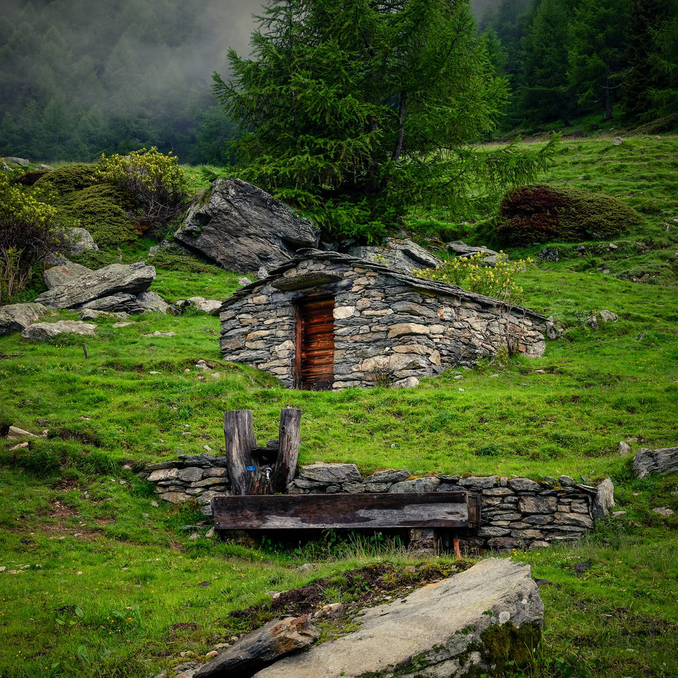 Petite cabane en pierre entourée de verdure avec des arbres et des collines en arrière-plan.