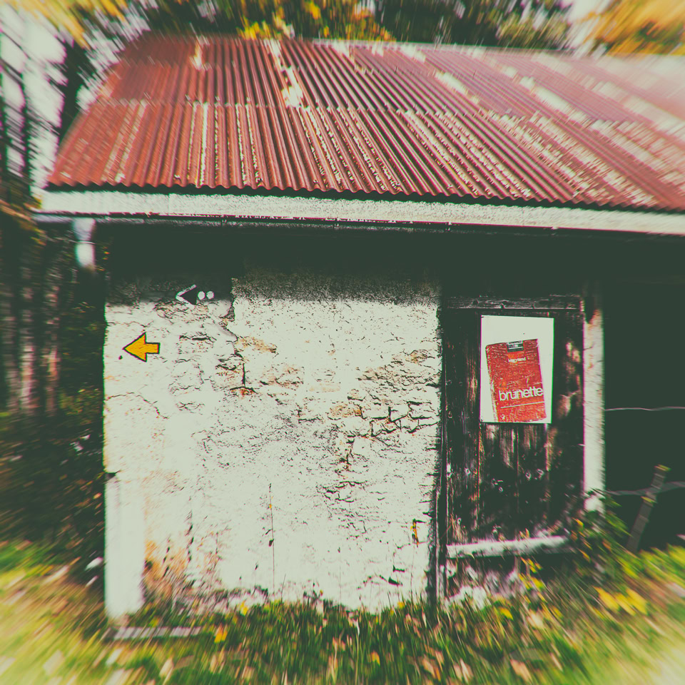 Petit bâtiment en bois avec un toit en tôle rouillée, une porte en bois avec une affiche rouge, et un mur blanc usé avec un panneau jaune.