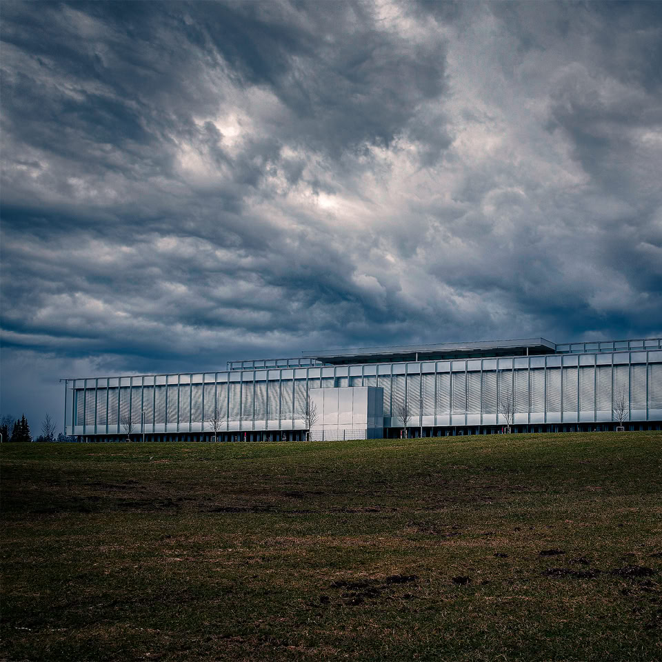 A large, modern glass building on a grassy field under a cloudy, dramatic sky.