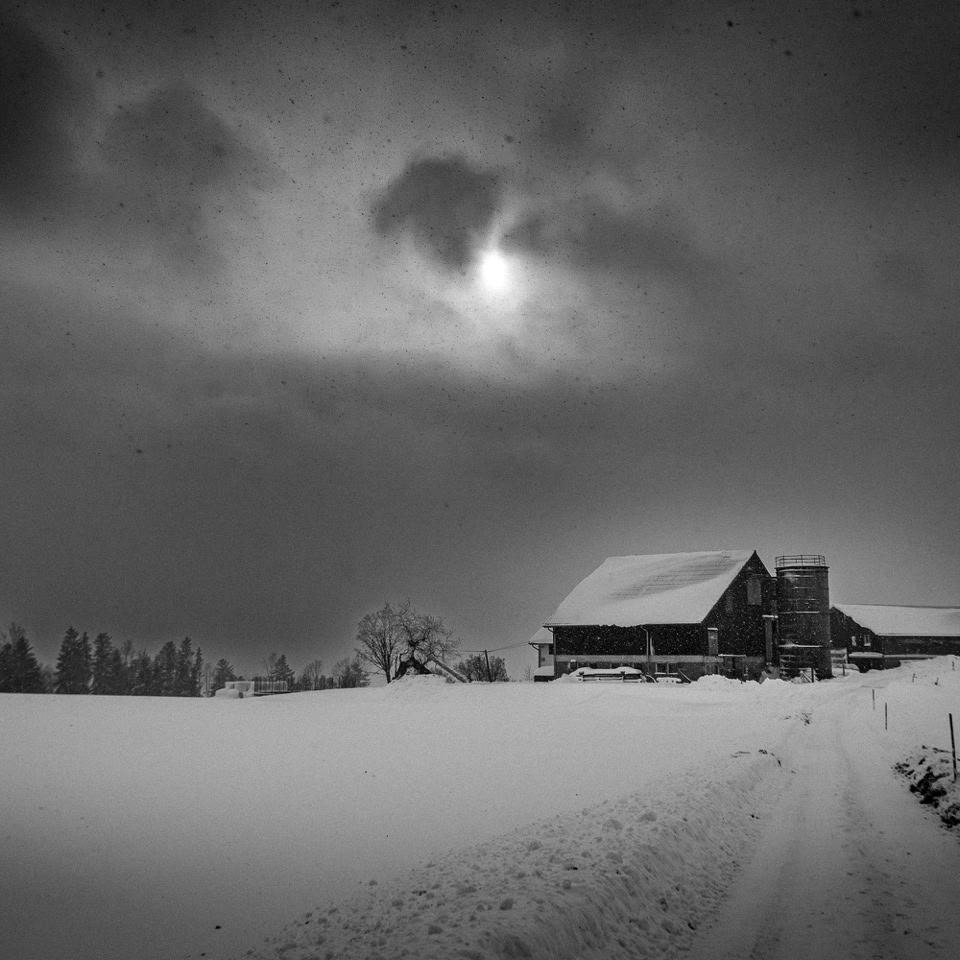 Paysage enneigé en noir et blanc avec une maison en bois, un ciel nuageux et un chemin menant à la maison.