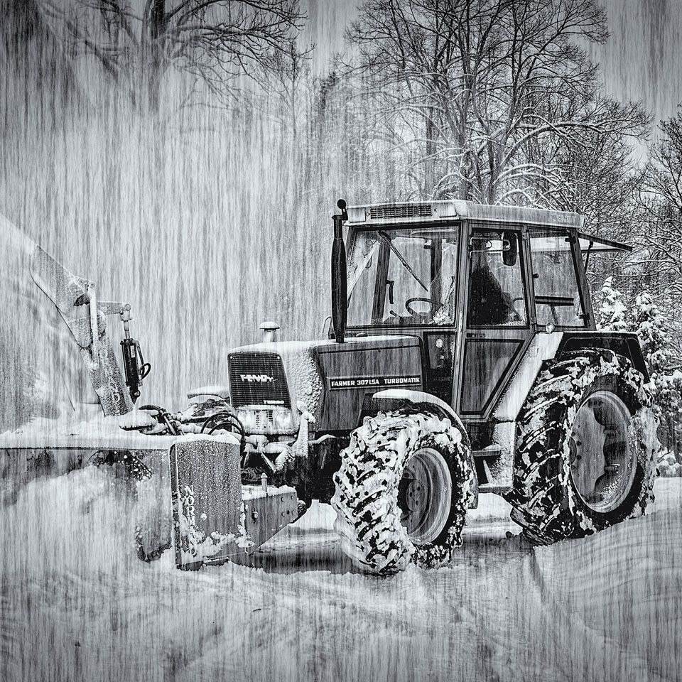Tracteur déneigeant une route enneigée sous la neige, avec des arbres en arrière-plan.