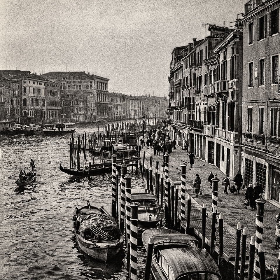 Vue en noir et blanc d'un canal avec des bateaux amarrés, des bâtiments anciens sur la droite et des personnes marchant le long du quai.