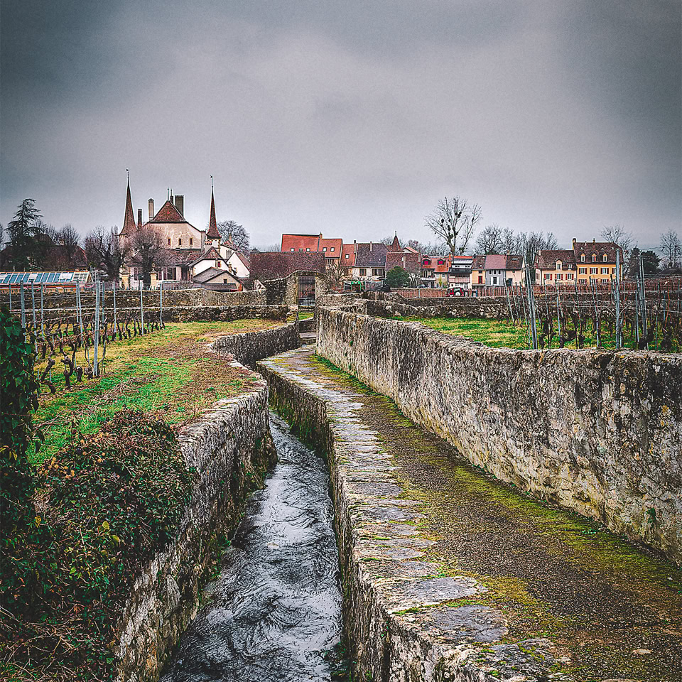 Un petit canal bordé de murs en pierre traverse un paysage rural avec des bâtiments et une église à l'arrière-plan sous un ciel nuageux.