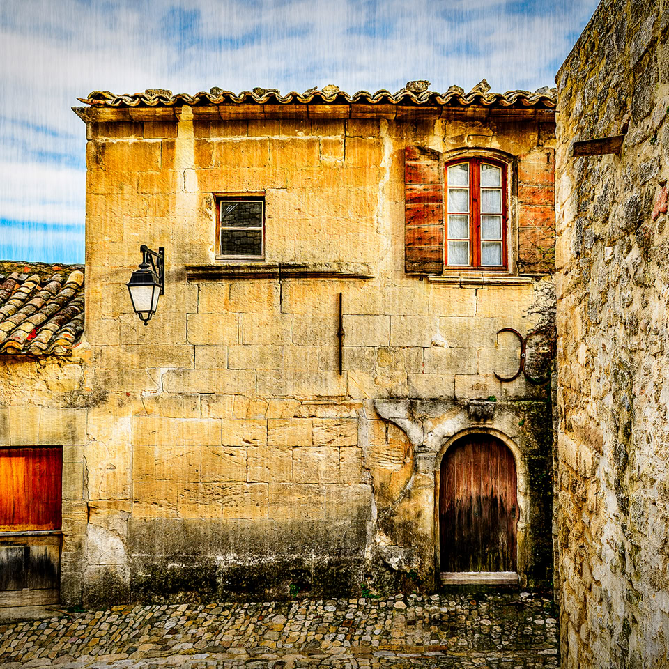 Vieille maison en pierre avec deux portes en bois et une fenêtre, entourée de murs en pierre sous un ciel partiellement nuageux.