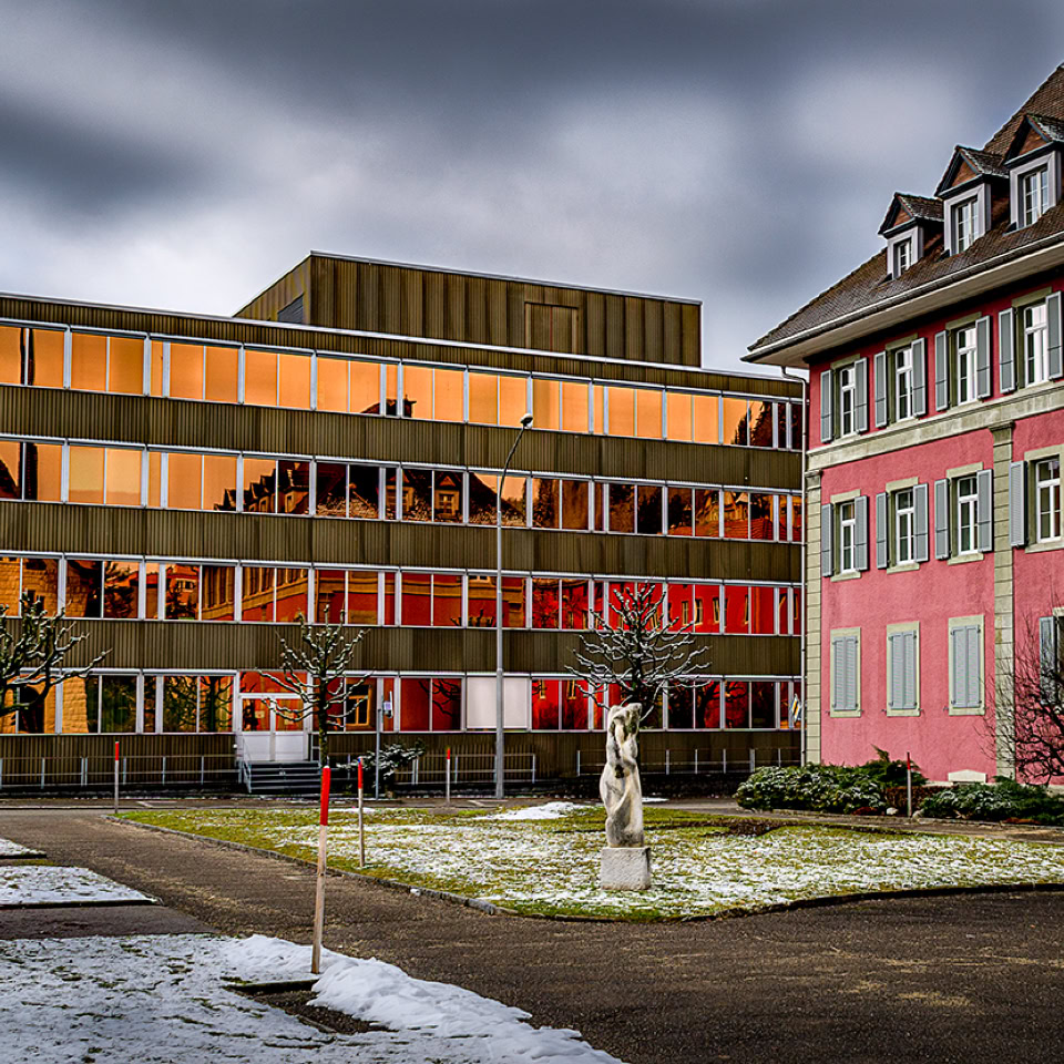 Bâtiments modernes et anciens côte à côte, avec des fenêtres illuminées, un ciel nuageux et un sol partiellement enneigé.