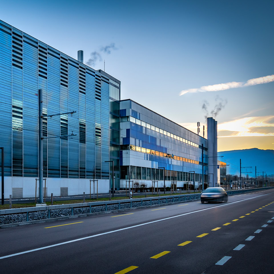 A modern glass building with reflective windows next to a road with a car driving by, under a clear sky.