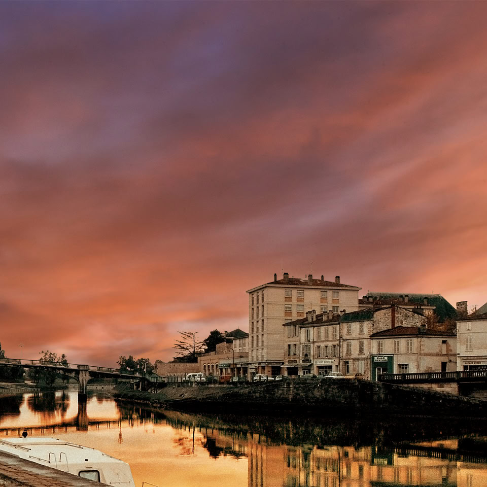 Vue d'un port avec des bâtiments historiques au bord de l'eau, sous un ciel de coucher de soleil aux teintes orange et rose.