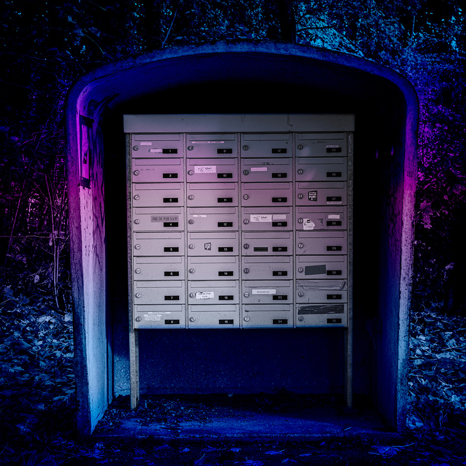 A row of mailboxes inside a small shelter, illuminated by blue and pink lights, with a dark forest background.